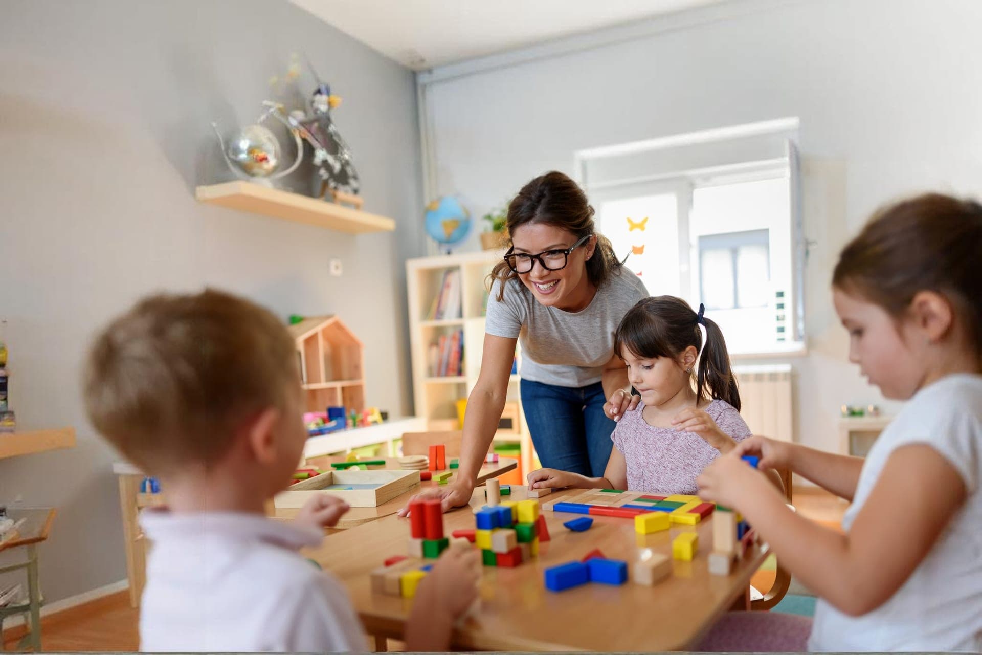 A smiling teacher helping children build with colorful wooden blocks in a bright classroom